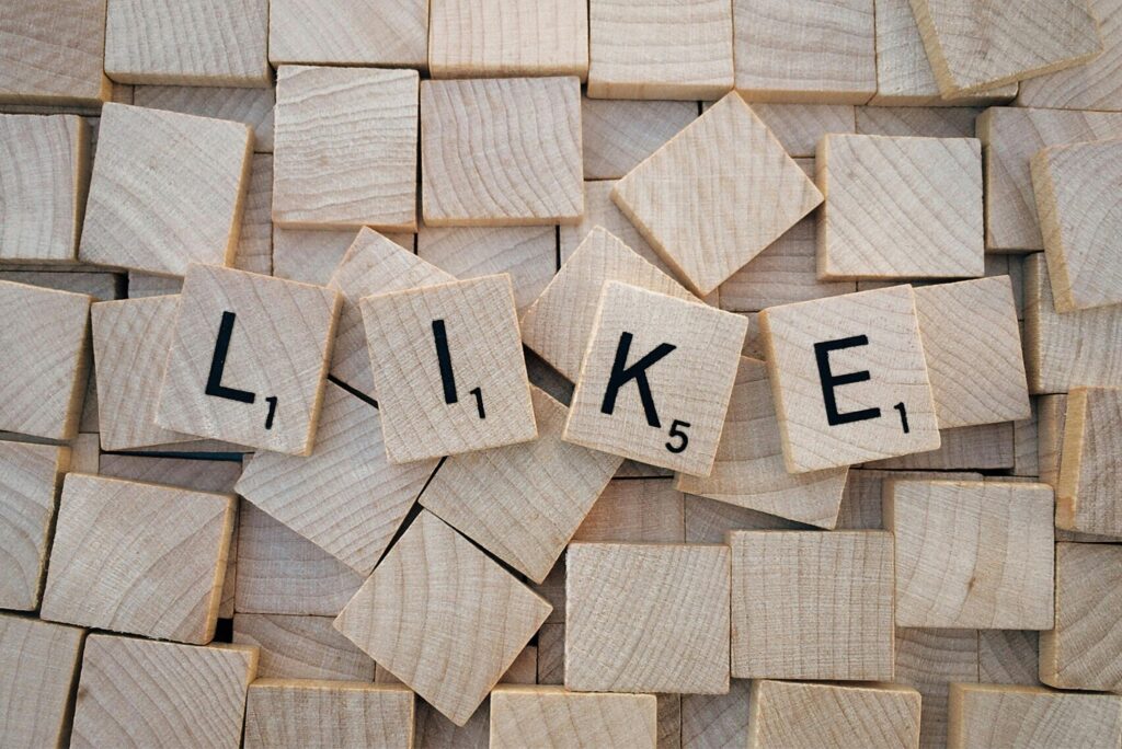 Top-down view of Scrabble tiles spelling 'LIKE' on a pile of blank wooden tiles.